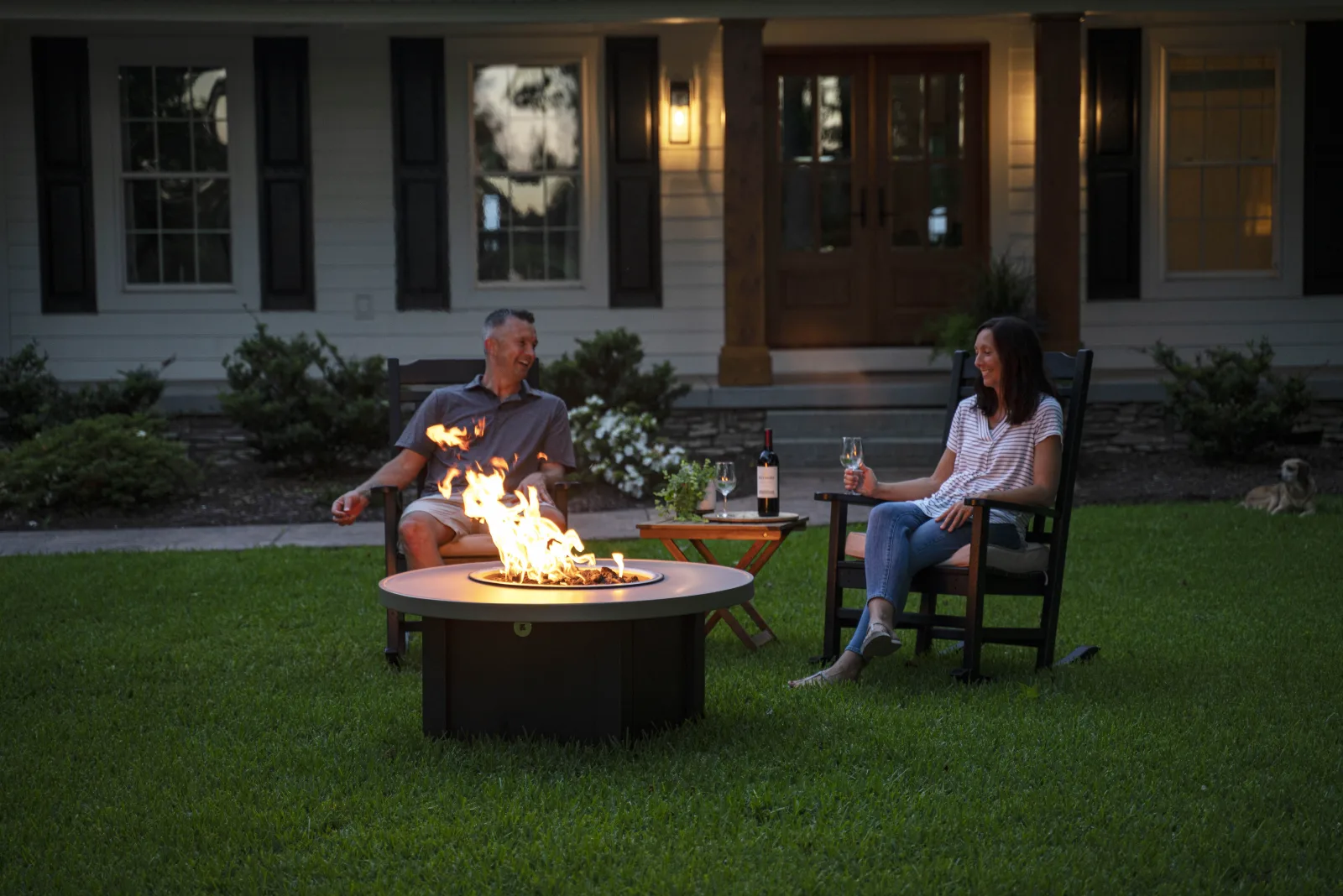 A couple in rocking chairs beside a Stoll fire pit on the lawn at dusk