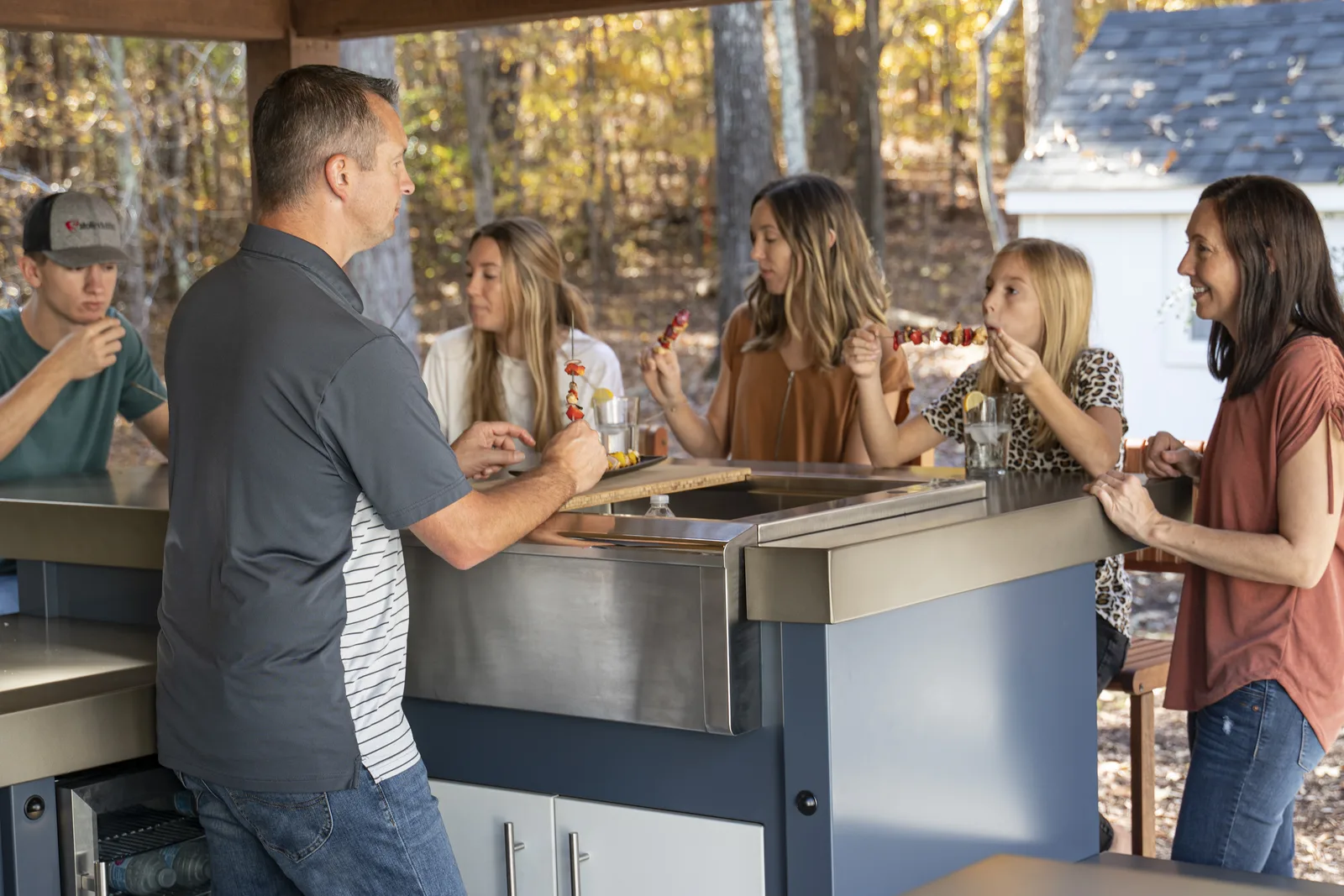 A family sharing kabobs around a Stoll outdoor kitchen island under a pergola